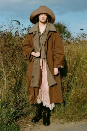 Model wearing a wide-brimmed hat, oversized brown coat, and boots standing in an overgrown field under a blue sky.