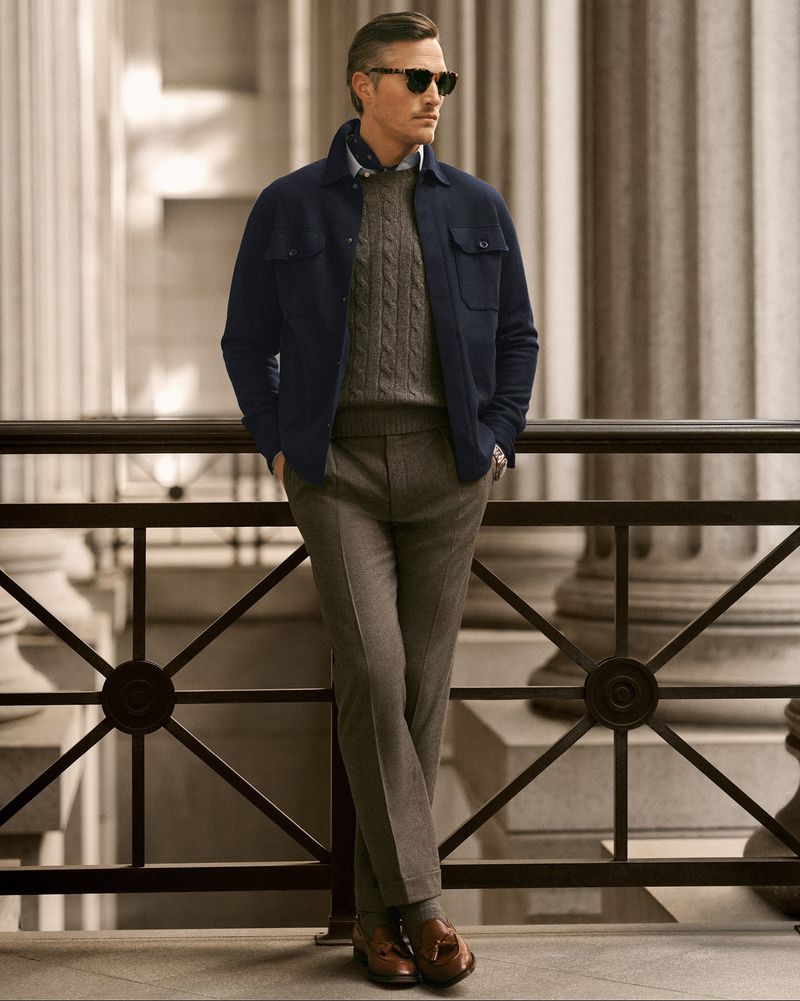 Adult man in navy wool jacket, grey sweater, and trousers stands outdoors against architectural backdrop in a fashion portrait.