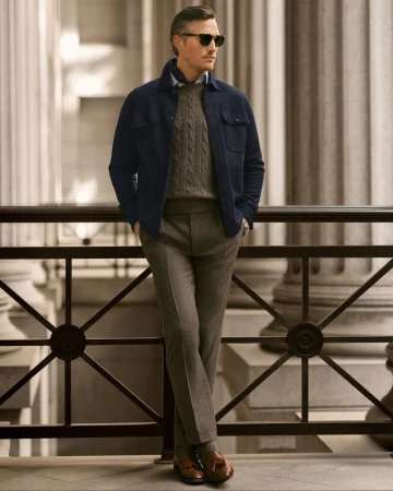 Adult man in navy wool jacket, grey sweater, and trousers stands outdoors against architectural backdrop in a fashion portrait.