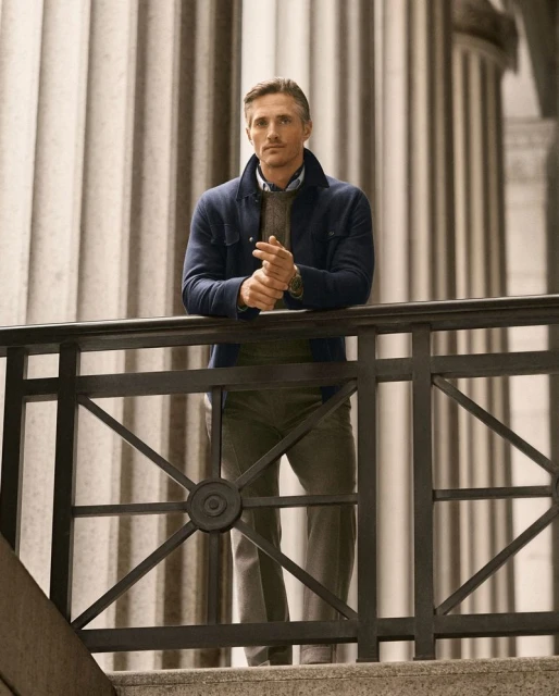 Man in blue jacket poses against marble columns and decorative railing in an outdoor setting with natural light, looking ahead. 