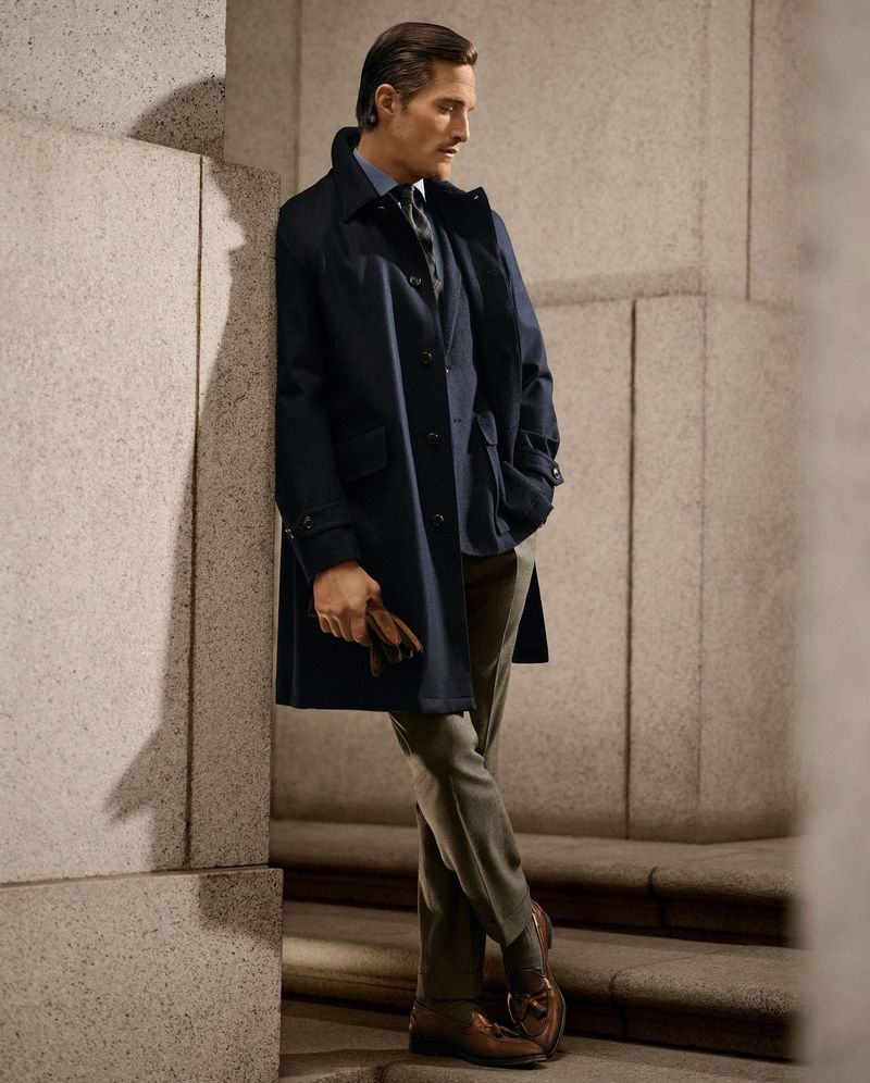 Man in navy overcoat leaning against column and steps. He has olive pants, tan loafers, and is holding gloves. He has dark hair.