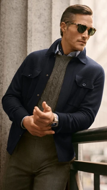 Male fashion shot: man in navy jacket, cable-knit sweater, grey trousers, and sunglasses poses against a stone column outdoors.