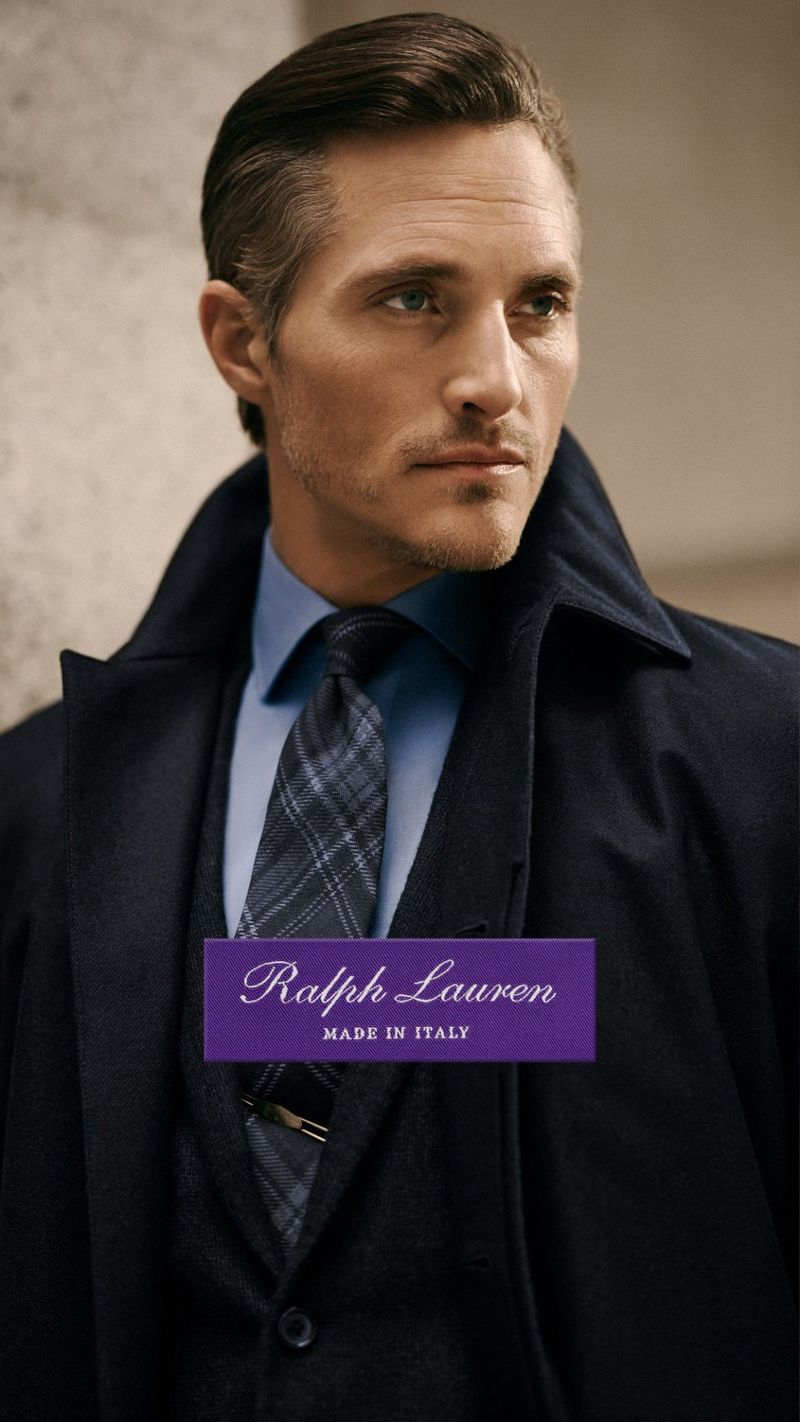 Close-up studio shot of a male model in a blue shirt, patterned tie, and dark coat, posing against a neutral wall. Fashion advertisement.