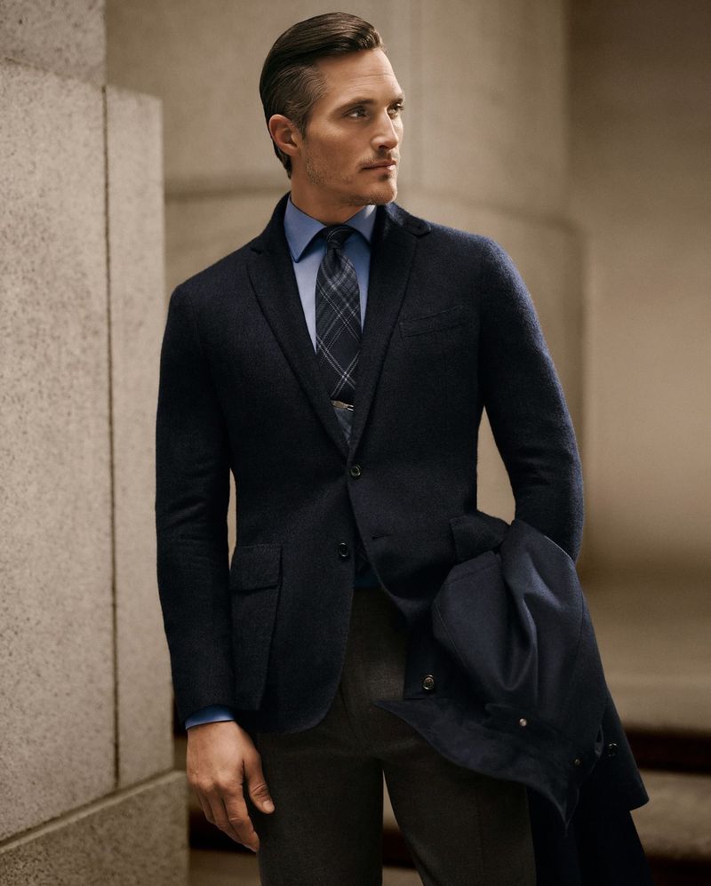 Man in dark blazer, blue dress shirt and plaid tie. He's holding a coat in his arm. He is in an urban outdoor environment.