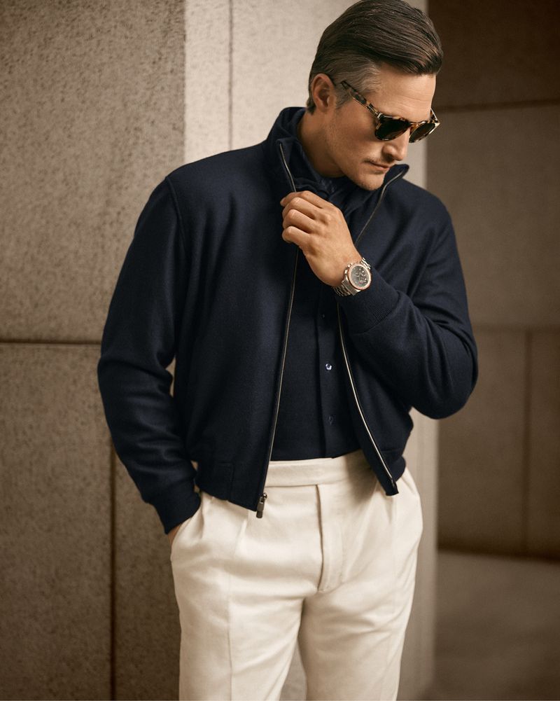 Mature man posing in a navy bomber jacket with a black shirt, cream-colored trousers and sunglasses in front of a textured wall.