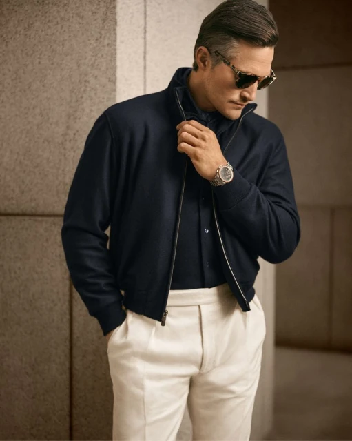 Mature man posing in a navy bomber jacket with a black shirt, cream-colored trousers and sunglasses in front of a textured wall.