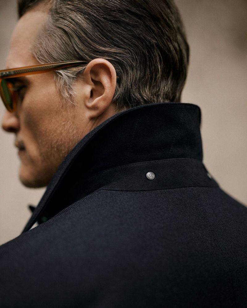 Adult male from behind wearing sunglasses and a dark overcoat with a high collar in a close studio shot with shallow depth of field.