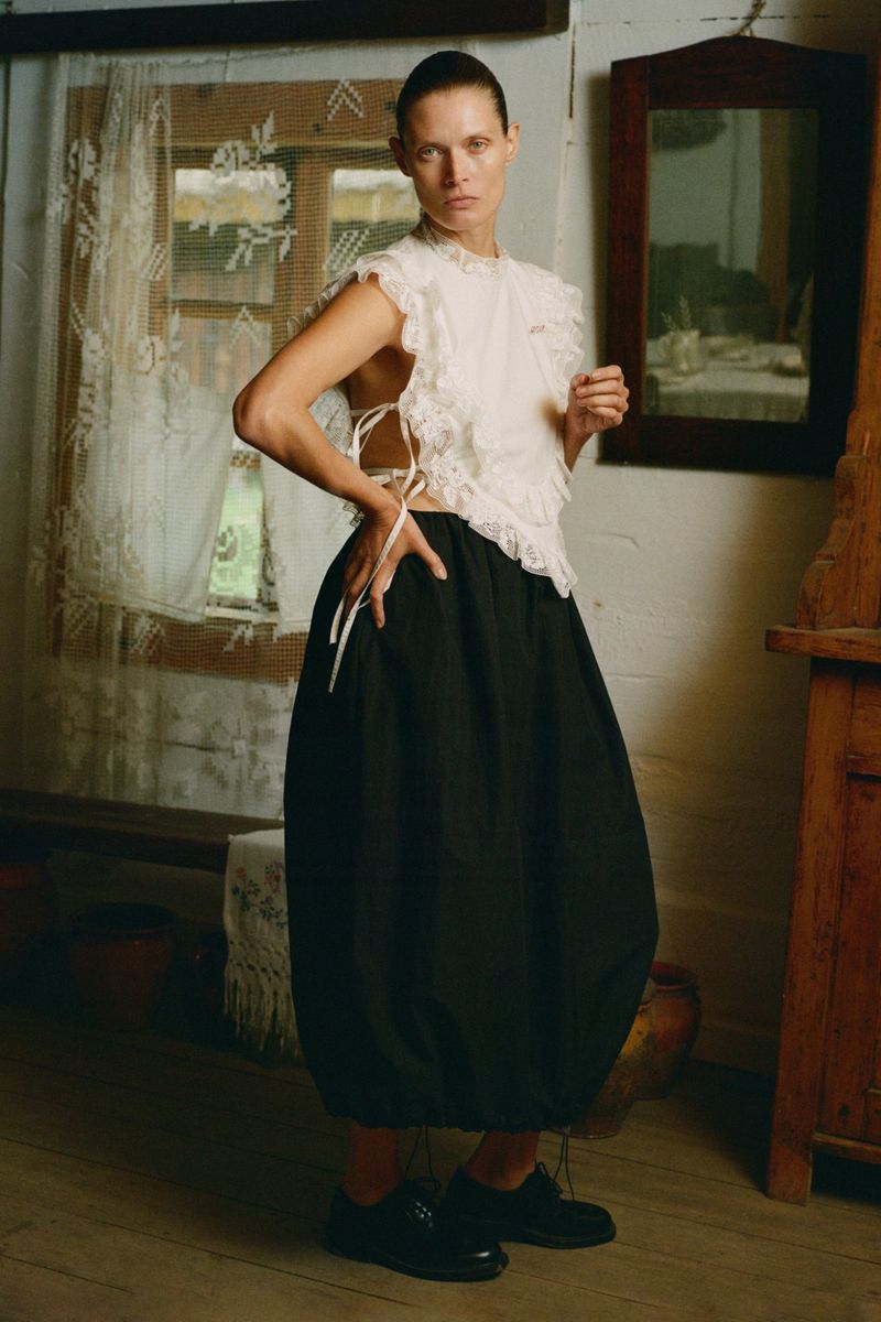 Fashion shot of a woman posing indoors wearing a white lace top, black skirt, and black shoes in a rustic setting.