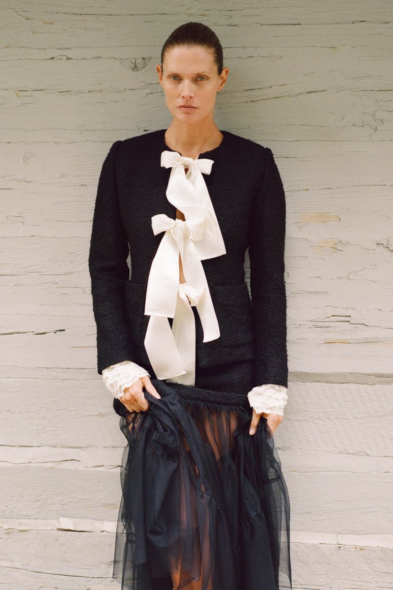 A stylish adult woman poses in a black jacket with a white ribbon and black tulle skirt against a weathered wood background.