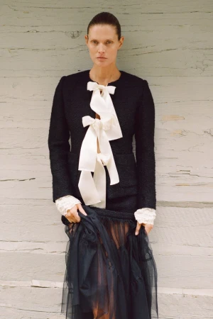 A stylish adult woman poses in a black jacket with a white ribbon and black tulle skirt against a weathered wood background.