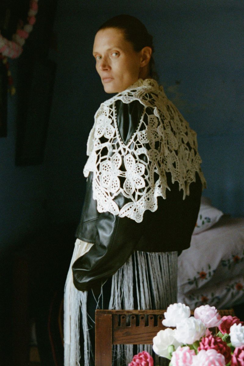 Adult woman in an indoor environment wearing a black leather jacket with a white lace shawl, looking toward the camera.