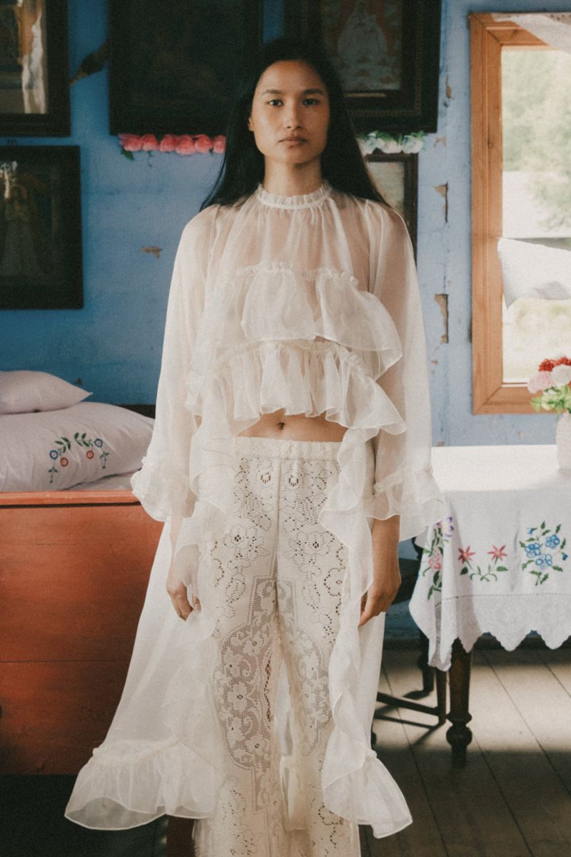A person stands indoors wearing white, ruffled clothing, looking at the camera. The indoor setting features rustic blue walls and decor.