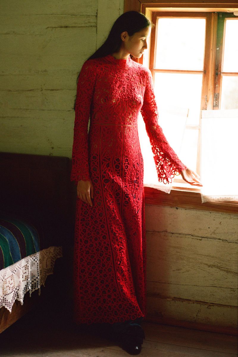 Young woman in long red lace dress poses near a bright window inside a rustic wooden building with a bed visible in the background.