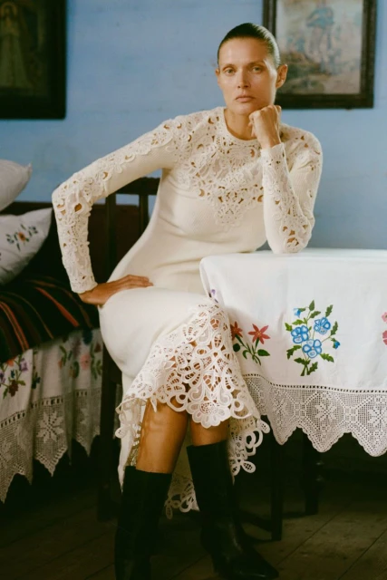 A fair-skinned woman in a long-sleeved dress sits at a table with an embroidered cloth, against a blue wall with framed art.