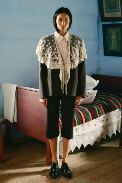A woman models a formal blazer with lace shawl, paired with black culottes and shoes, set against a muted blue interior with folk art.