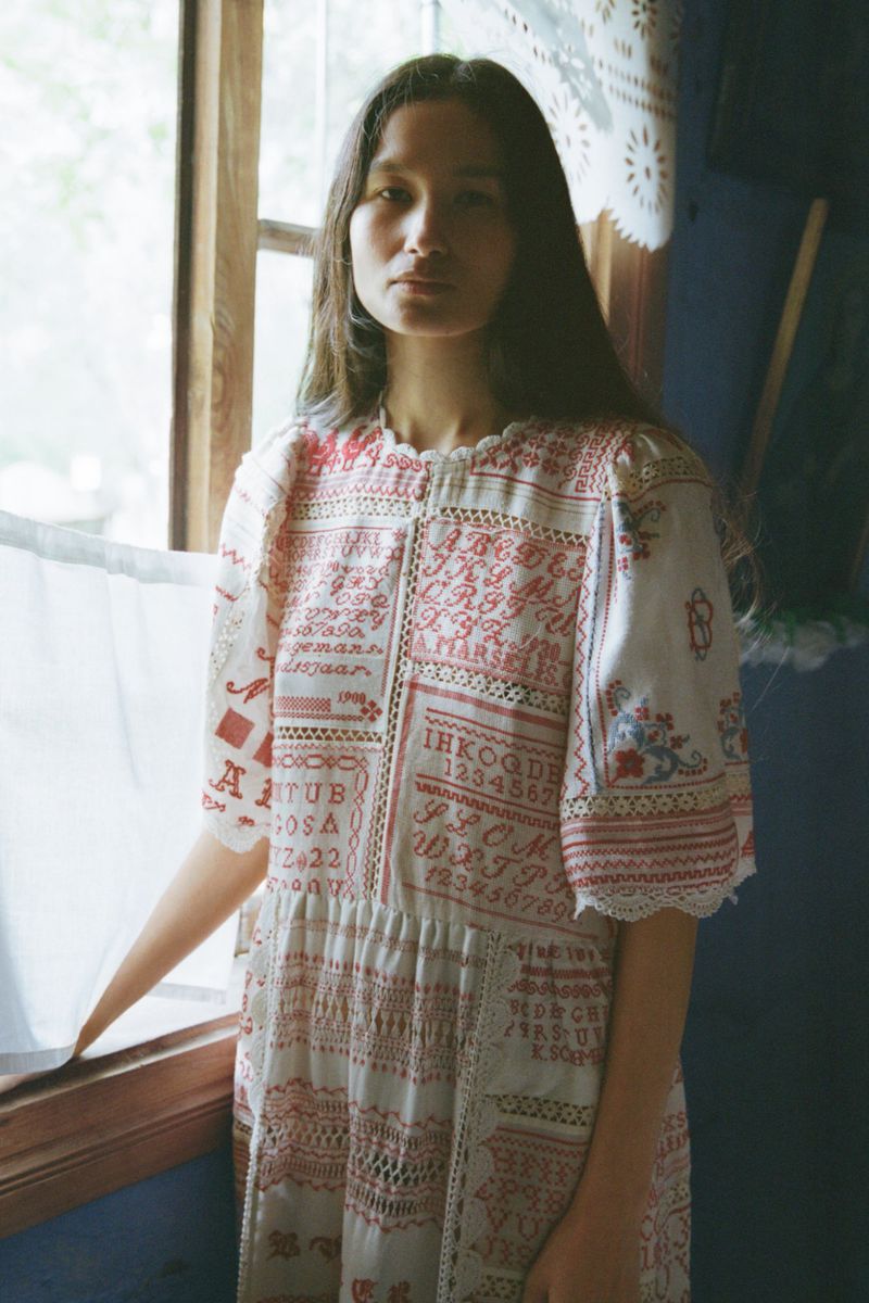 Model standing indoors near a wooden window wearing a white patterned dress with red embroidery details.