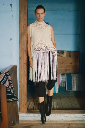 A woman poses in a doorway wearing a white lace sleeveless top with fringe, a black skirt, and black boots inside a rustic home.