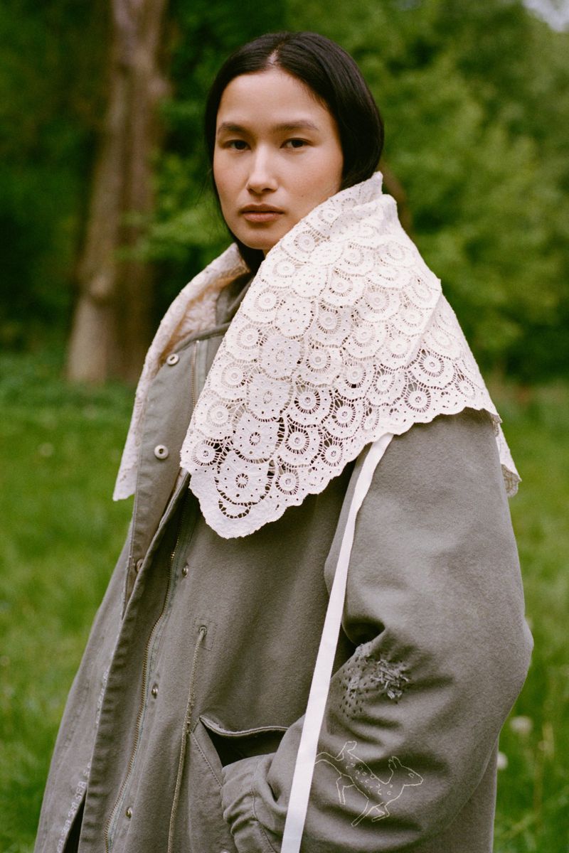 Adult female poses outdoors wearing a grey-green coat with a decorative lace collar standing in a blurred meadow environment.