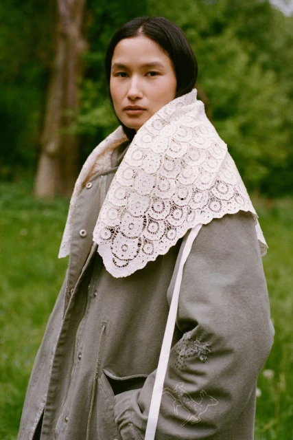 Adult female poses outdoors wearing a grey-green coat with a decorative lace collar standing in a blurred meadow environment.