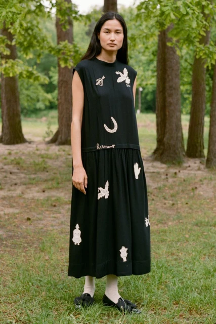 Adult female standing in a grassy area, wearing a black dress with white embroidery, outdoors, eye-level shot, nature background.