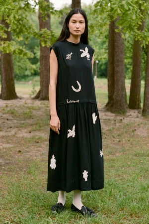 Model standing outdoors in a black embroidered midi dress wearing white socks and dark shoes against a wooded background