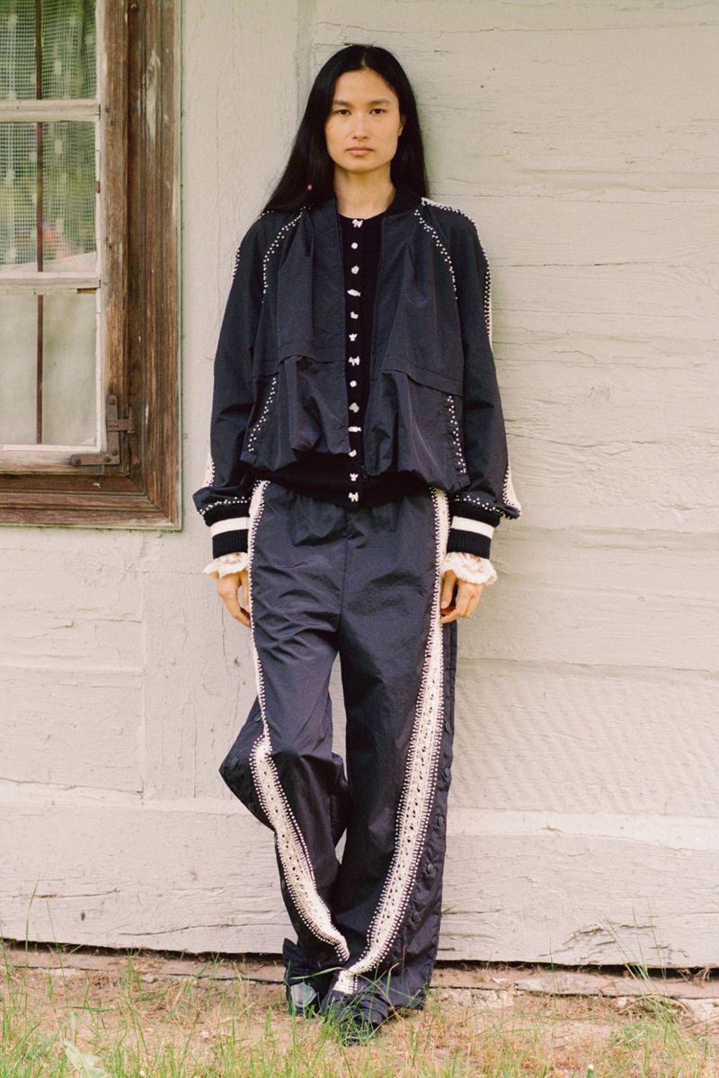 Portrait of a young woman wearing a black jacket and pants with white trim details standing in front of a weathered wooden wall.