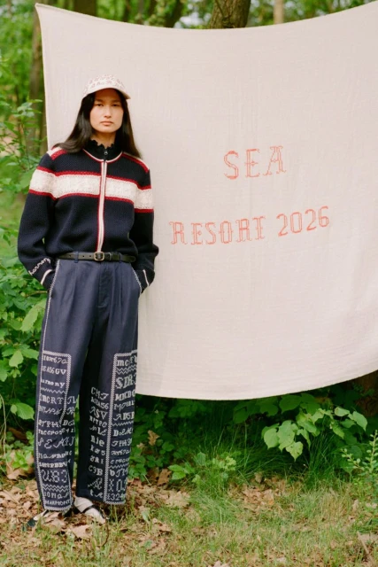 Woman wearing a patterned sweater and pants stands near a white backdrop with 'SEA Resort 2026' text in a natural, outdoor setting.