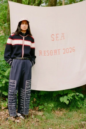 Woman wearing a patterned sweater and pants stands near a white backdrop with 'SEA Resort 2026' text in a natural, outdoor setting.