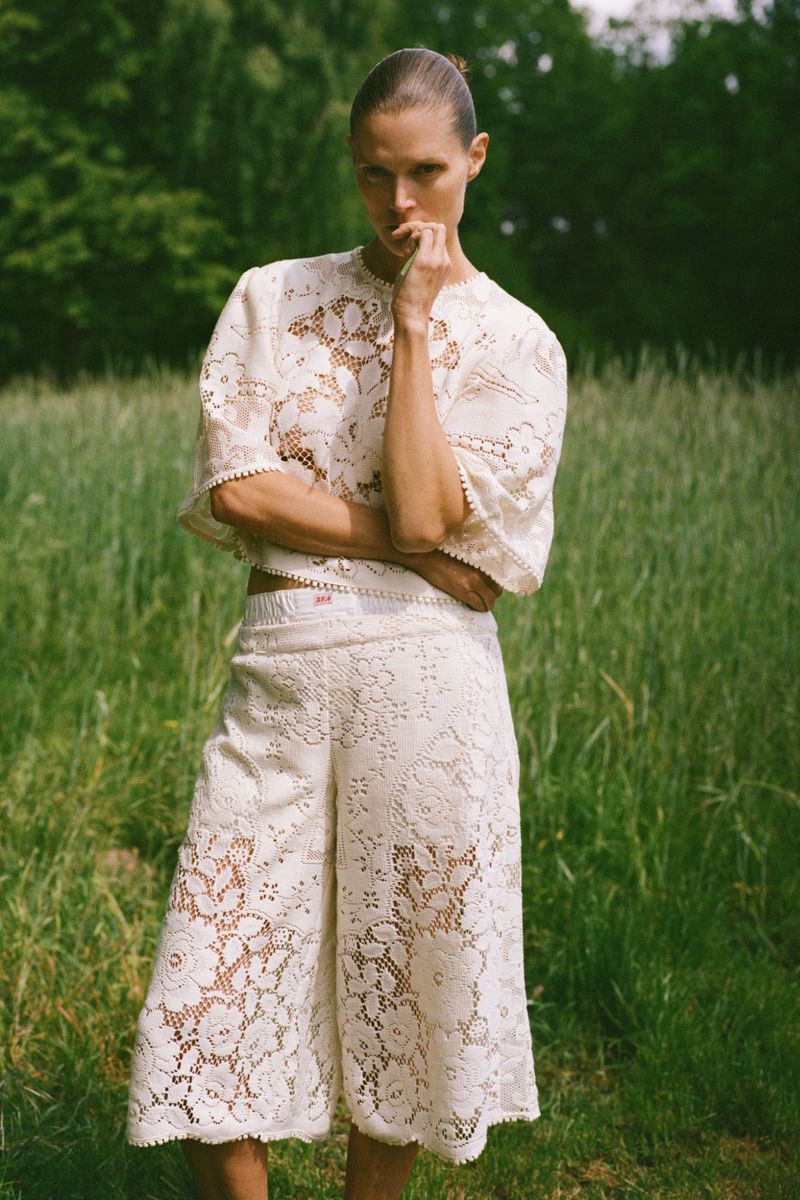 Adult female wearing a white lace top and culottes stands in a grassy field with trees. Fashion editorial shot outdoors.