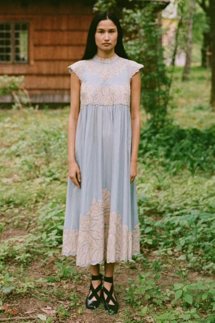 A full-length shot of an adult female posing outdoors in a light blue dress with embroidered floral patterns, set against a rustic background.