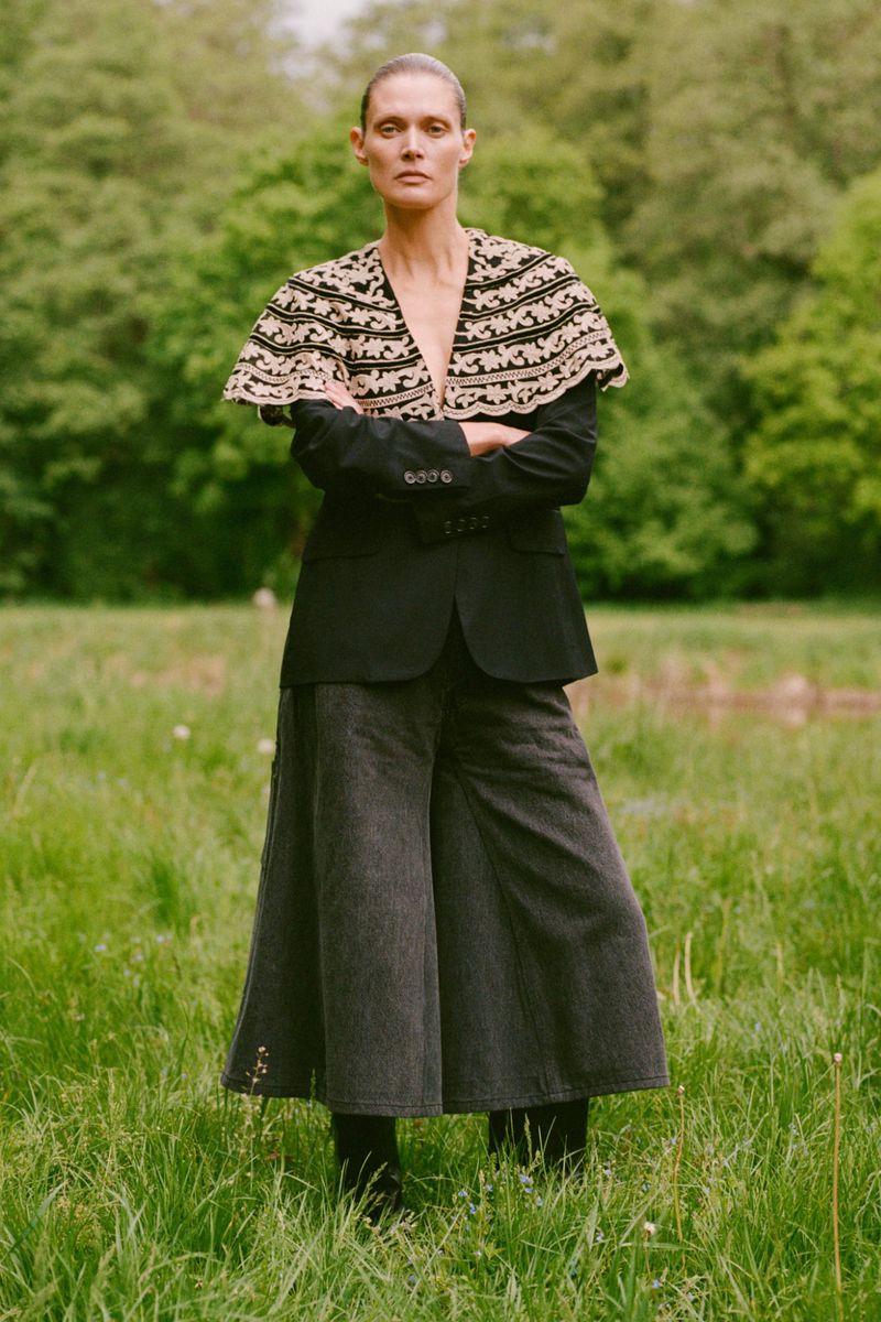 Model Malgosia Bela wearing an embroidered cape and dark blazer standing in a grassy meadow for Sea RS26 Lookbook