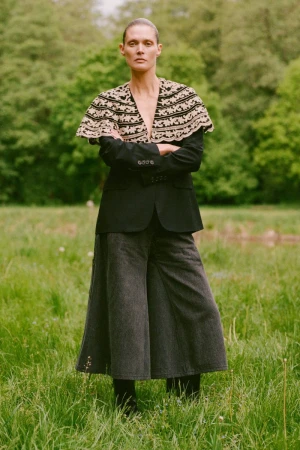 Model Malgosia Bela wearing an embroidered cape and dark blazer standing in a grassy meadow for Sea RS26 Lookbook