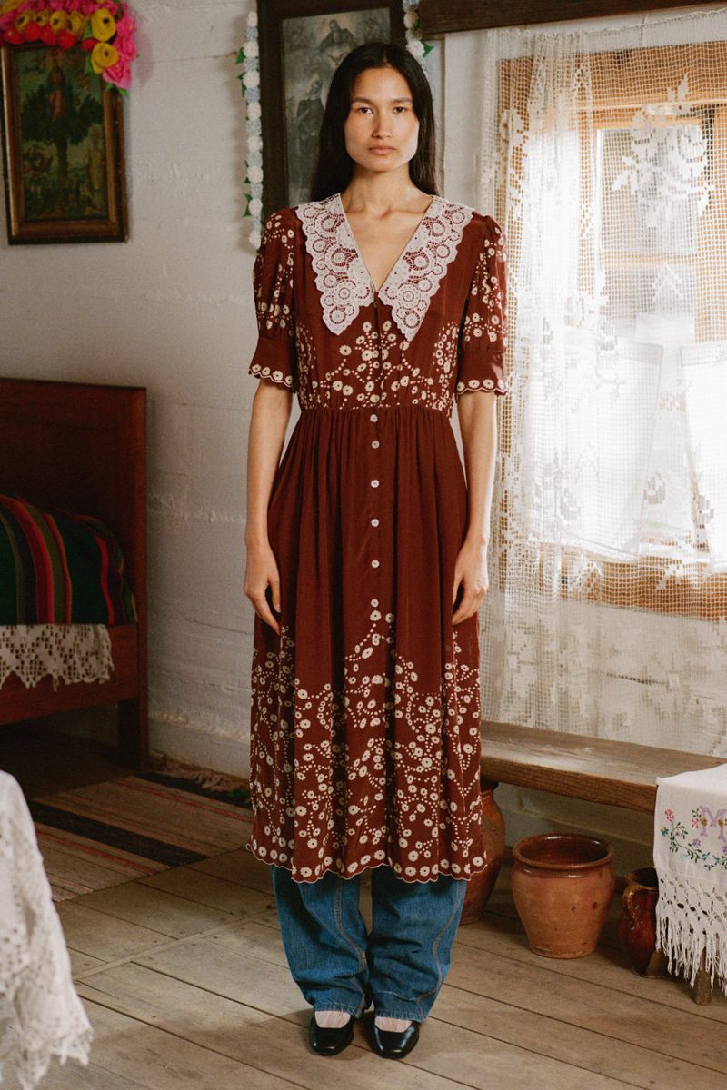 A woman in a rust-colored floral dress stands in a rustic room with a window, bed, and painted walls, with a neutral expression.
