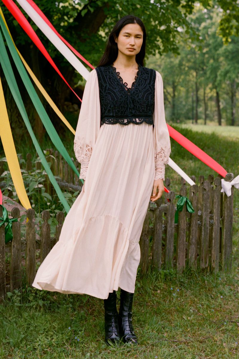 Woman wearing a long dress and boots standing in a grassy field with decorative ribbons on a fence in the background.