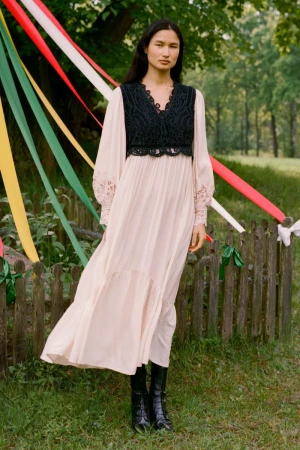 Woman wearing a long dress and boots standing in a grassy field with decorative ribbons on a fence in the background.