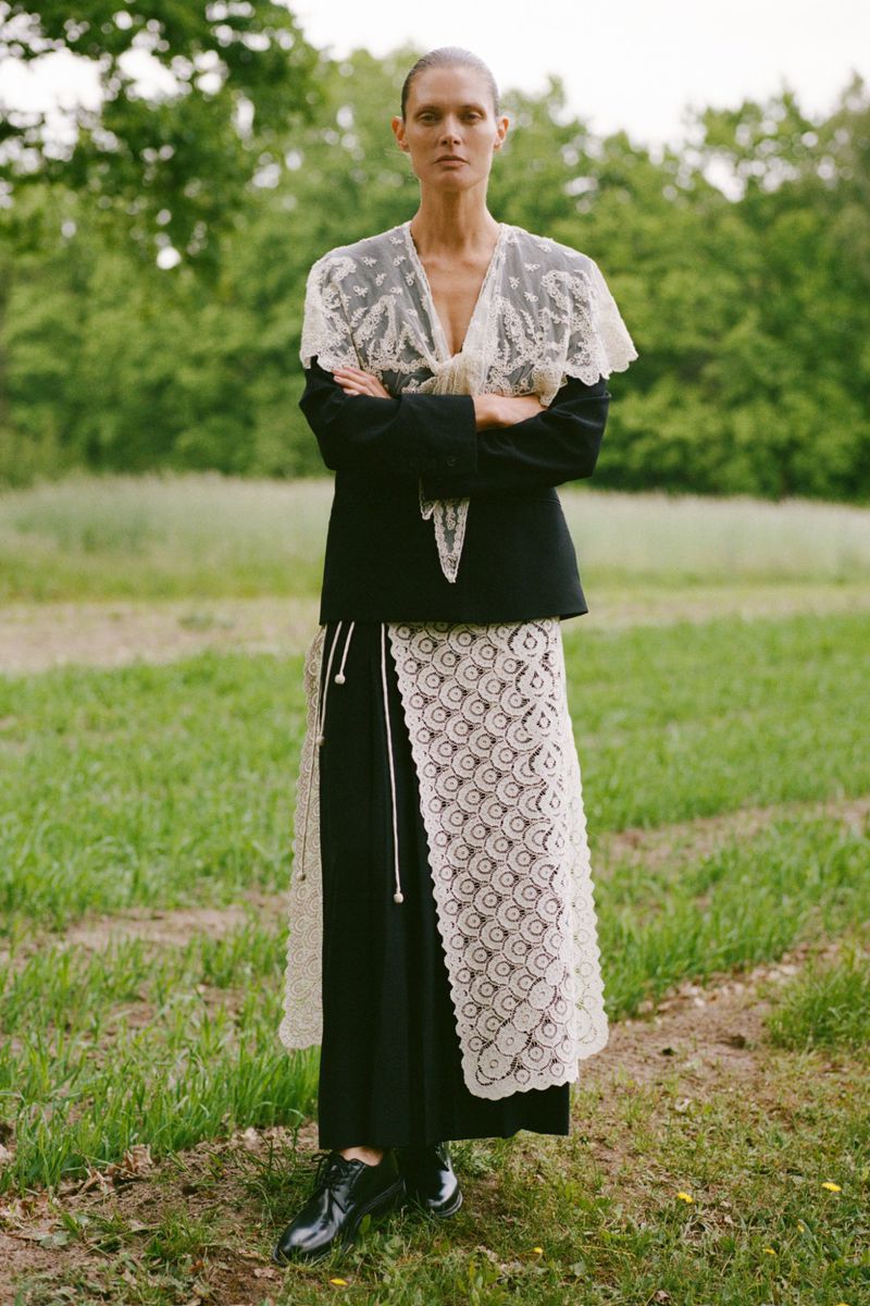An adult female in a black jacket with lace overlay, standing in a grassy field with trees, creating a striking fashion portrait.