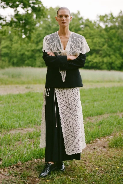 An adult female in a black jacket with lace overlay, standing in a grassy field with trees, creating a striking fashion portrait.