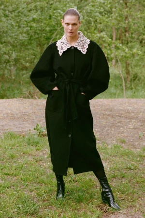 Model wearing a long black coat with a decorative white lace collar standing outdoors in a wooded setting.
