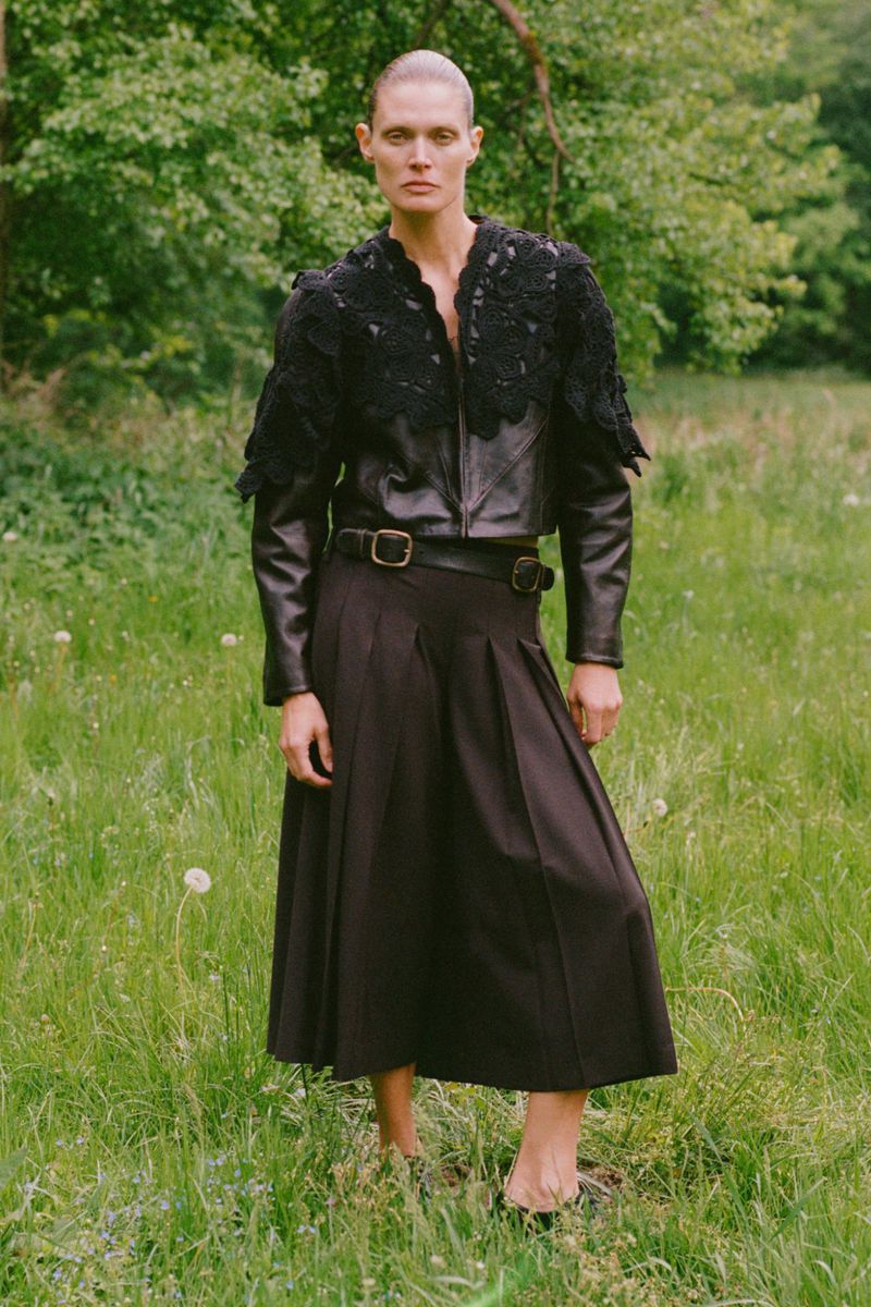 Woman in black leather jacket and dark pleated skirt stands in grassy field, trees in background, fashion editorial, outdoor setting.