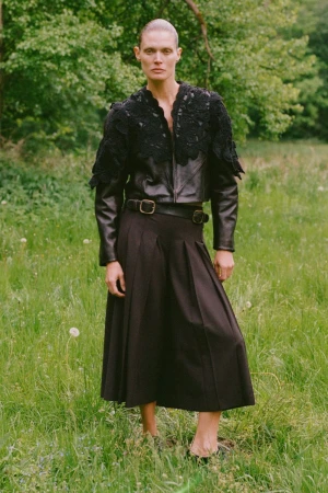 Woman in black leather jacket and dark pleated skirt stands in grassy field, trees in background, fashion editorial, outdoor setting.