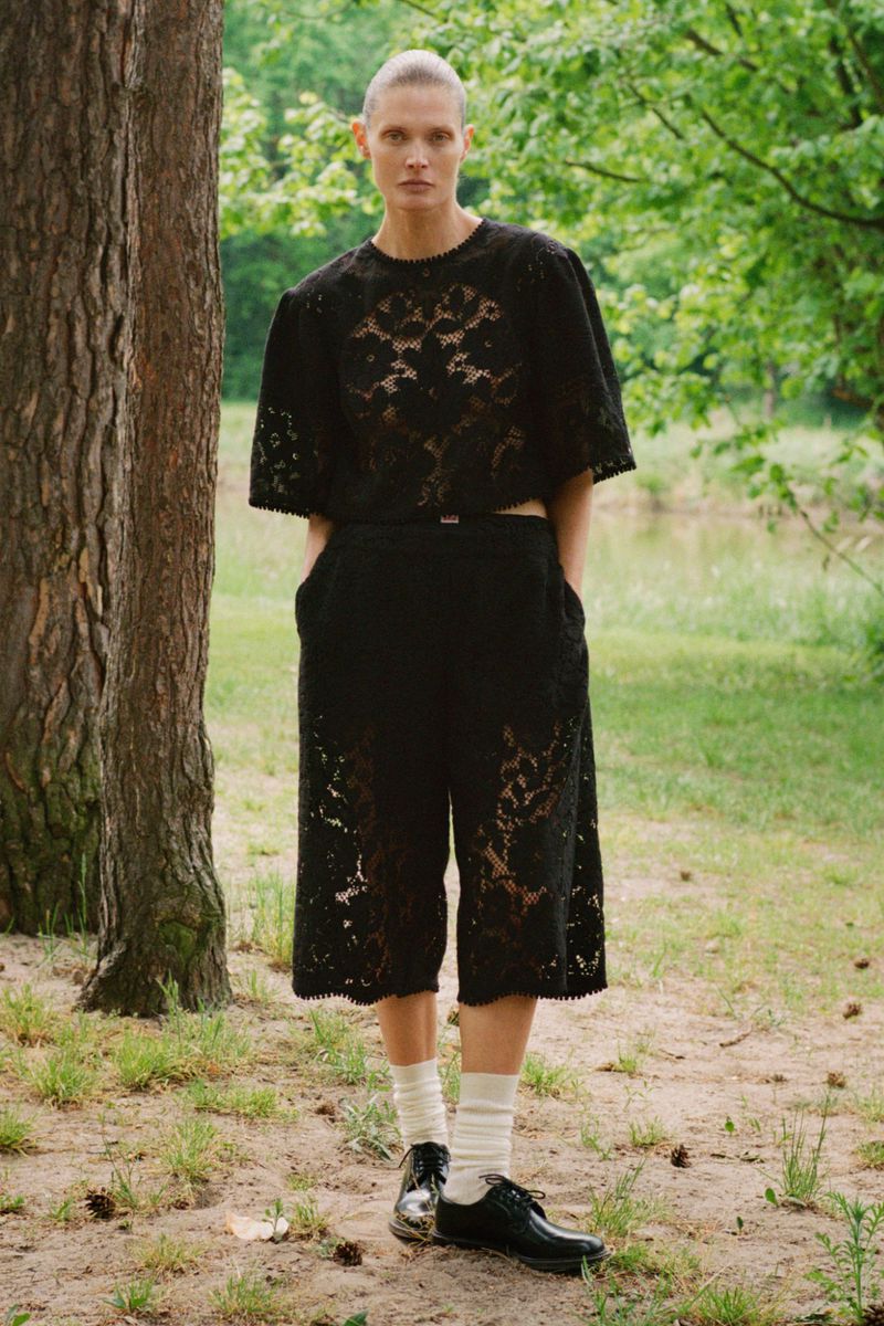 Model Malgosia Bela wearing a black lace short-sleeve top and matching culottes outdoors by trees.