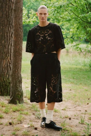Model Malgosia Bela wearing a black lace short-sleeve top and matching culottes outdoors by trees.