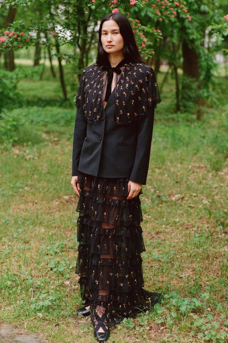 Woman in floral cape and tiered dress stands in forest.  Black blazer, black shoes, floral pattern, greenery. Fashion editorial, outdoor shot.