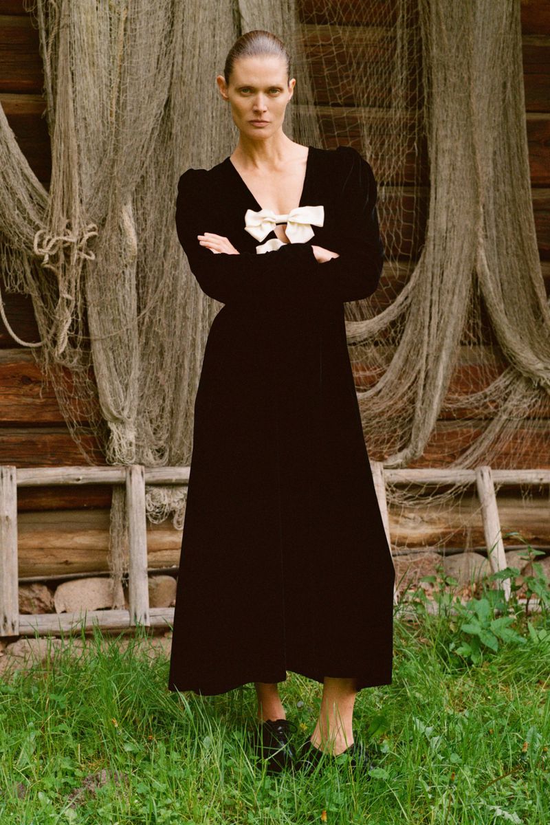 Adult woman in a black dress with a white bow poses in front of a weathered building and fishing net. Fashion editorial lifestyle photography.