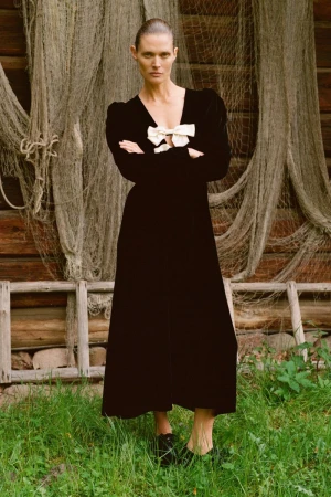 Adult woman in a black dress with a white bow poses in front of a weathered building and fishing net. Fashion editorial lifestyle photography.