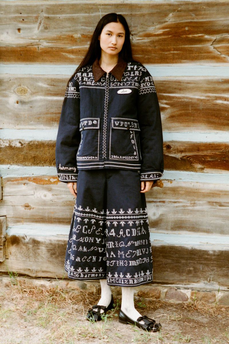A woman in an embroidered jacket and skirt poses in front of a wooden wall, creating a fashion editorial shot with natural outdoor light.