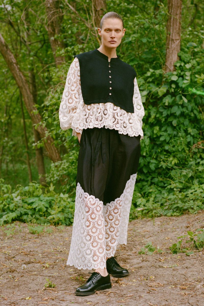 Full-shot of a woman with a shaved head wearing a black and white lace dress with black shoes, standing outdoors in a forest-like environment.
