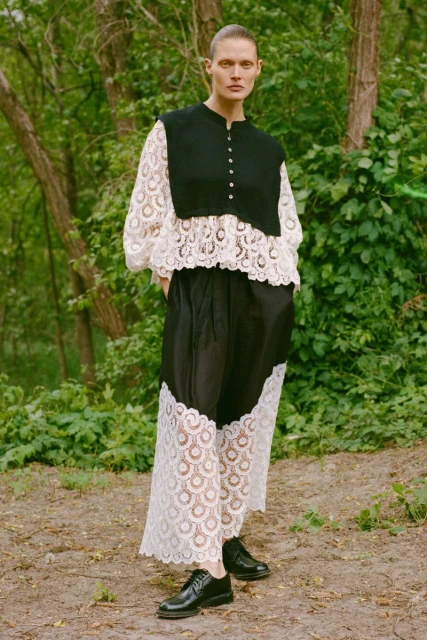Full-shot of a woman with a shaved head wearing a black and white lace dress with black shoes, standing outdoors in a forest-like environment.