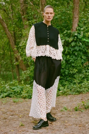 Full-shot of a woman with a shaved head wearing a black and white lace dress with black shoes, standing outdoors in a forest-like environment.