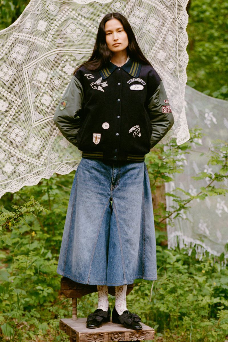 Full-length portrait of a woman wearing a varsity jacket and long denim skirt posing on a wooden stool in an outdoor wooded setting.